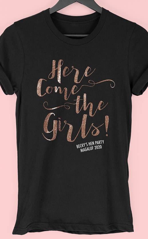 Image to buy product Here Come The Girls Glitter Personalised Hen Party T Shirt. Script lettering in glitter rose gold print with subtext in white on a black t-shirt.