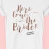 Image to buy product Here Comes The Bride Glitter Personalised Hen Party T Shirt. A bride's version of the design in script lettering in glitter rose gold print with subtext in black on a white t-shirt.